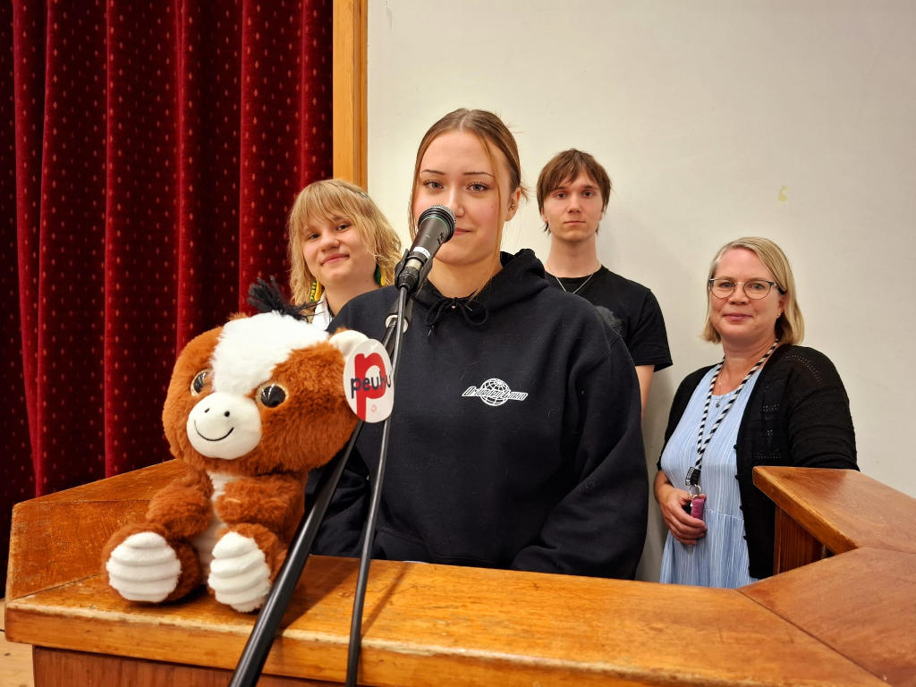 Kolme opiskelijaa ja yksi opettaja seisovat puhujanpöntössä mikrofonin takana.
